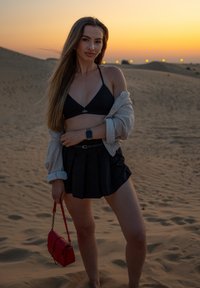 Melnas bikini top, pelēks pogājams krekls un melna izliekta svārki. Tur rokās mazu sarkanu rokassomu, stāv uz smiltīm ar saulrieta fonu.