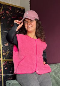 Femme souriante, portant un gilet polaire rose, un chemisier à manches longues noir, un pantalon gris et une casquette de baseball rose, assise sur une chaise verte texturée.
