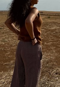 Top senza maniche a maglia marrone abbinato a pantaloni a palazzo a righe con vita alta. Sullo sfondo si vede un terreno secco simile alla paglia.