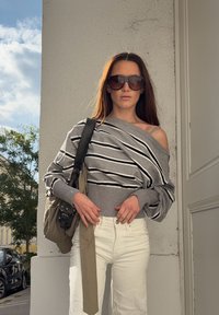 Egy nő oversized szürke csíkos pulóvert visel, amely az egyik válláról lóg, fehér nadrágot, nagy napszemüveget �és egy bézs táskát hordoz a szabadban, egy épület közelében.