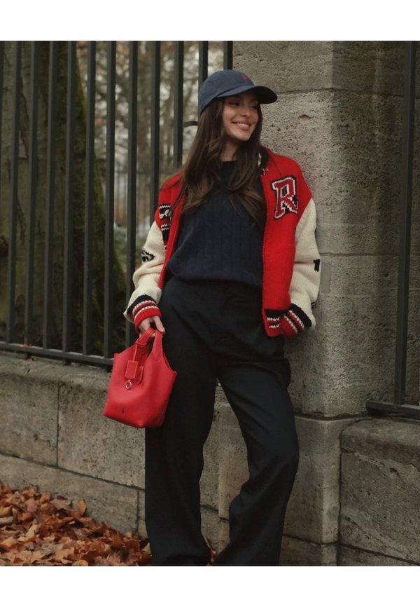 Red and cream varsity jacket with black stripes, navy jumper, wide-legged black trousers, and a small red handbag. Background with stone wall and leaves.