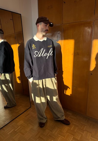 Hombre con sudadera oscura con texto "Aloft", pantalones anchos beige, gorra marrón, de pie junto a un armario de madera y un espejo de cuerpo entero bajo la luz del sol.
