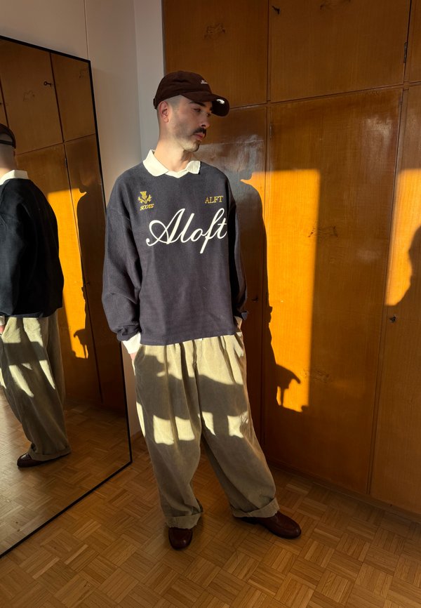 Mann trägt dunklen Pullover mit "Aloft"-Schriftzug, beige weite Hose, braune Mütze, steht bei einem Holzkleiderschrank und Ganzkörperspiegel im Sonnenlicht.