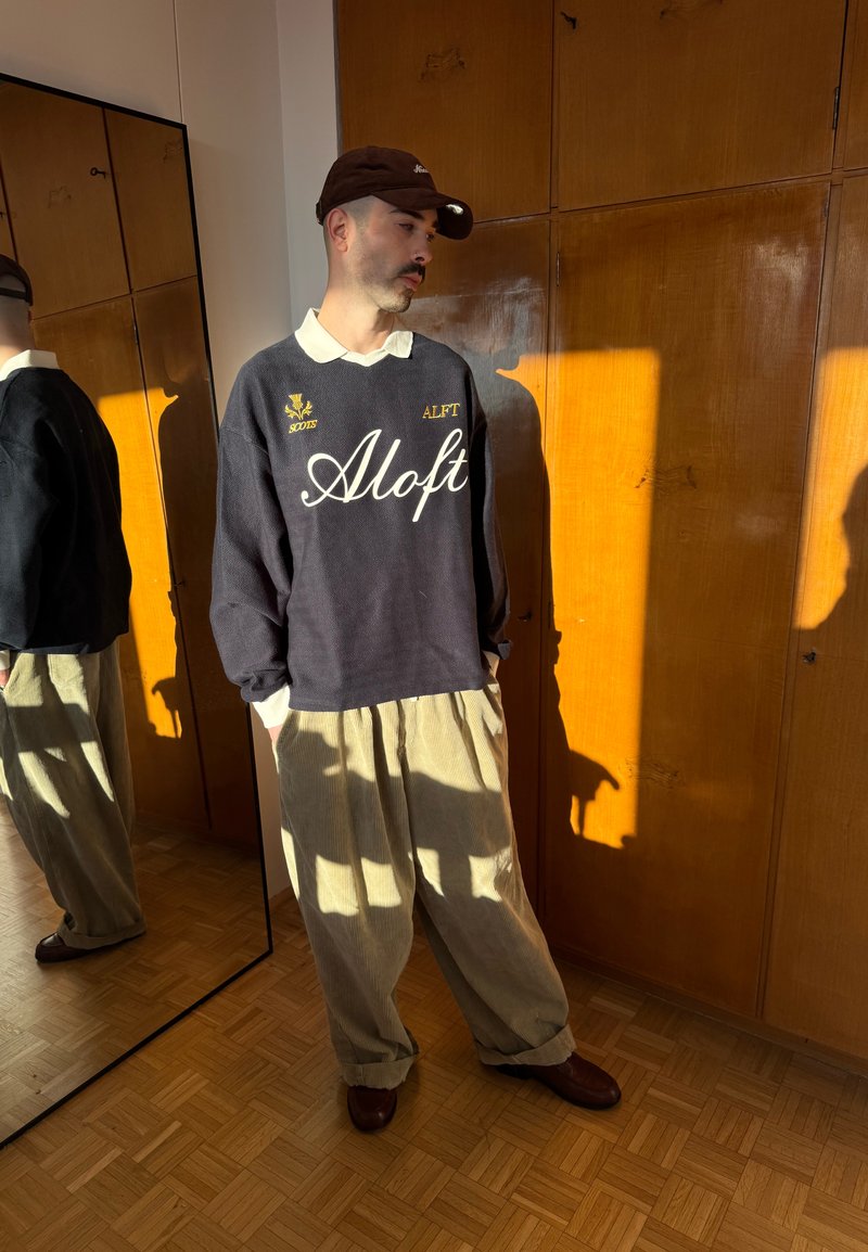 Hombre con sudadera oscura con texto "Aloft", pantalones anchos beige, gorra marrón, de pie junto a un armario de madera y un espejo de cuerpo entero bajo la luz del sol.