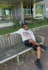 Felpa grigia casual con la scritta "ACADEMY", pantaloni corti neri e scarpe da ginnastica nere. Seduto su una panchina di legno consumata circondata da fiori.