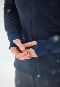Manteau bleu marine texturé avec fermeture éclair ; les poignets ajustés couvrent les mains. Le tissu est lisse, avec un design en couches et une coupe près du corps.
