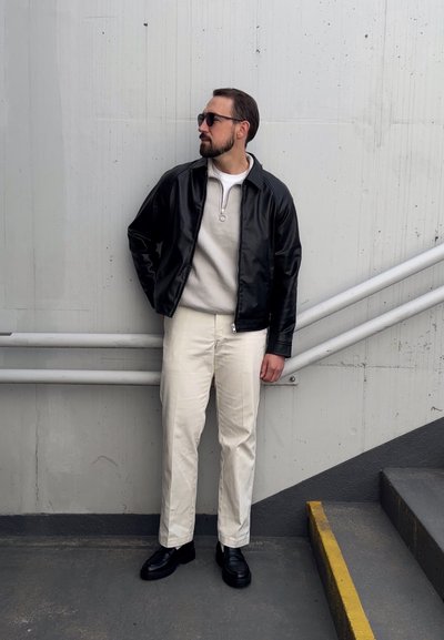 Chaqueta de cuero negra sobre un sudadera gris claro, pantalones blancos y zapatos negros. El fondo presenta una pared gris y barandillas blancas.