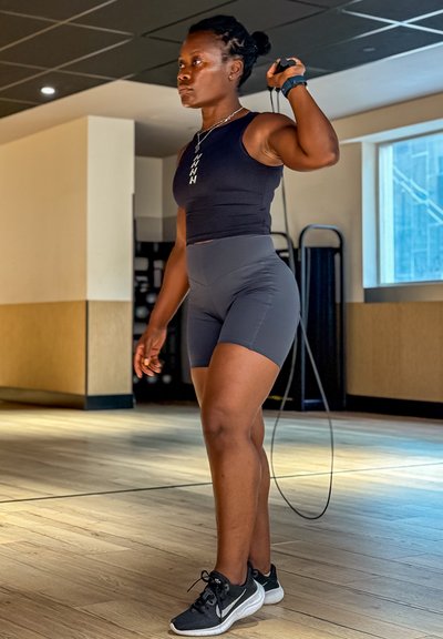 Top deportivo negro y pantalones cortos grises en una persona que sostiene una cuerda para saltar. Zapatillas negras; ambiente interior con suelo de madera y luz de ventana.