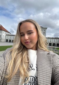 Vaaleat hiukset, yllään ruudullinen beige bleiseri ja valkoinen neulepaita, jossa on teksti; taustalla pilvinen taivas ja historiallinen rakennus.