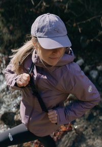 Giacca sportiva viola chiaro con un design aderente, cappuccio regolabile e logo. Berretto grigio con logo. Leggings scuri. Ambientazione all'aperto con rocce e acqua.