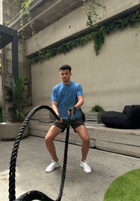 Melnas cīņu virves, kuras tur persona zilā krekliņā un šortos, sportojot uz betona grīdas ar zaļumiem un sēdvietām fona.