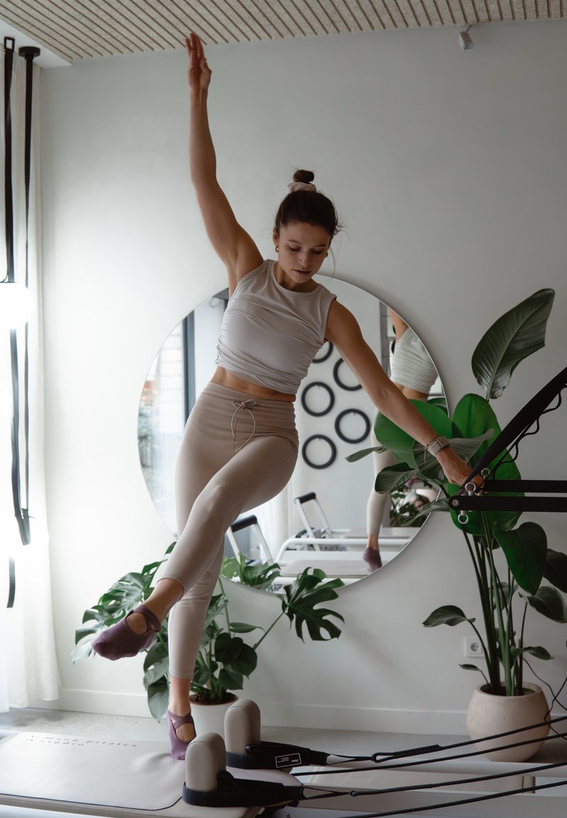 Réformateur de fitness avec une personne effectuant un exercice, portant un débardeur clair et des leggings beiges, avec des chaussettes violettes, entourée de plantes.