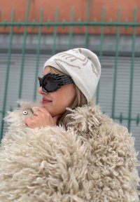Femme portant un bonnet en tricot crème, des lunettes de soleil futuristes noires et un manteau beige duveteux, debout à l'extérieur près d'une clôture métallique verte.