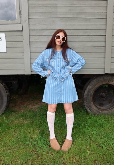 Mujer de pelo largo y grandes gafas de sol blancas de pie sobre el césped, vestida con un vestido azul a rayas, calcetines blancos hasta la rodilla y zapatos color beige.