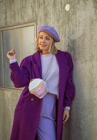 Μωβ μακρύ παλτό πάνω από έναν ανοιχτό μωβ πουλόβερ, συνδυασμένο με λεβάντα παντελόνι. Μια τσάντα σε σχήμα cupcakes σε παστέλ χρώματα κρέμεται από τον ώμο της.