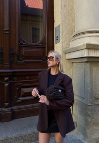 Rjav oversized blazer, črna obleka, teksturirana rjava torbica, velike črne sončne očala, stoji ob lesenih vratih z zapletenimi gravurami.