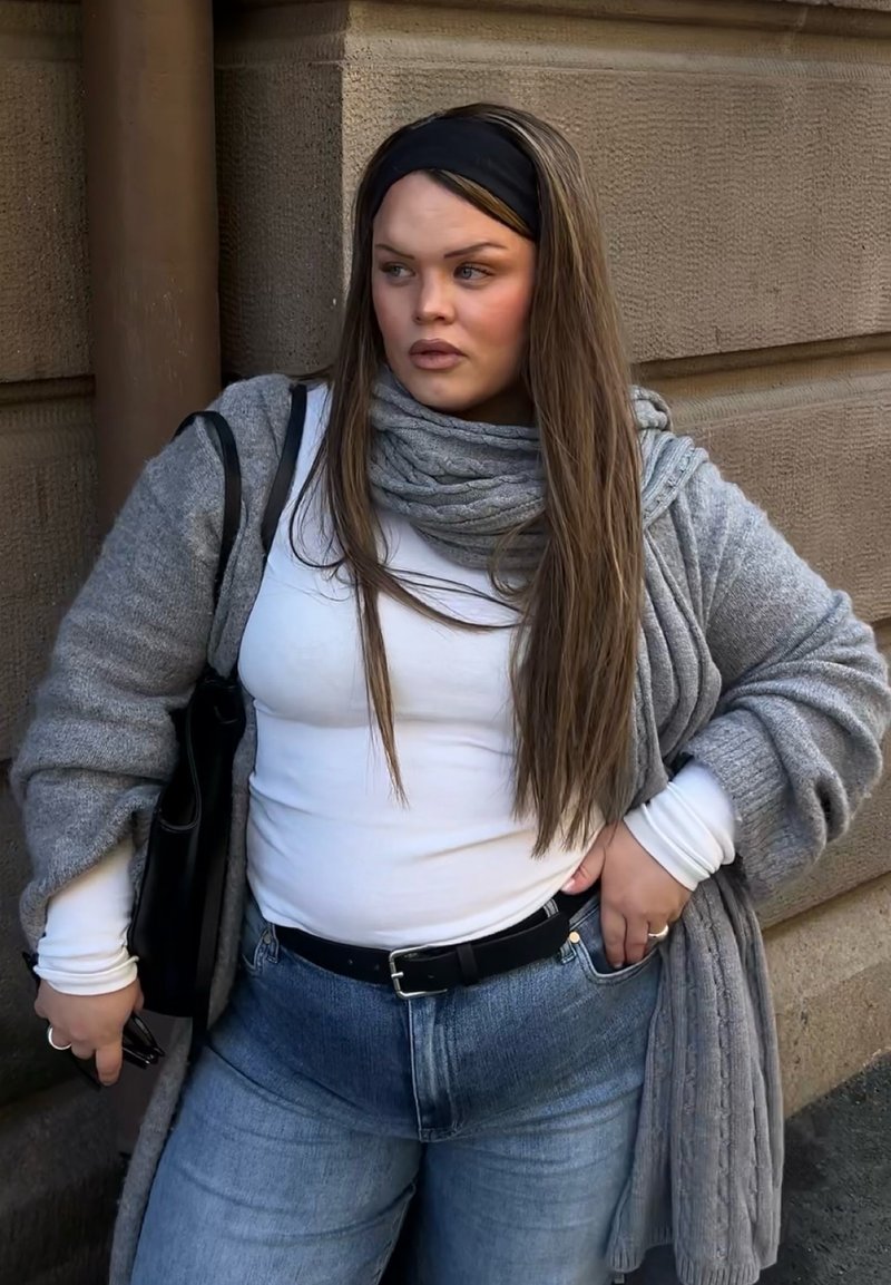 Frau mit langen Haaren und Stirnband, die ein weisses Top, eine graue Strickjacke, einen Schal und Jeans trägt, steht an einer Steinmauer, die Hand in der Tasche, hält eine Sonnenbrille.