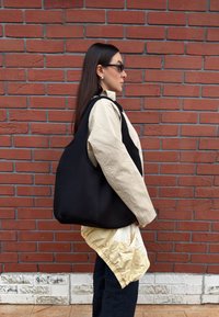 Femme aux longs cheveux noirs portant des lunettes de soleil, une veste beige, un sac noir sur l'épaule, debout de profil contre un mur de briques rouges.