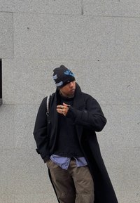 Μαύρο μακρύ παλτό πάνω από μια υφή μαύρη ζακέτα, γκρι καρό πουκάμισο και ελιές cargo παντελόνι. Μαύρο σκουφάκι με μπλε γράμματα. Αξεσουάρ περιλαμβάνουν δαχτυλίδια.