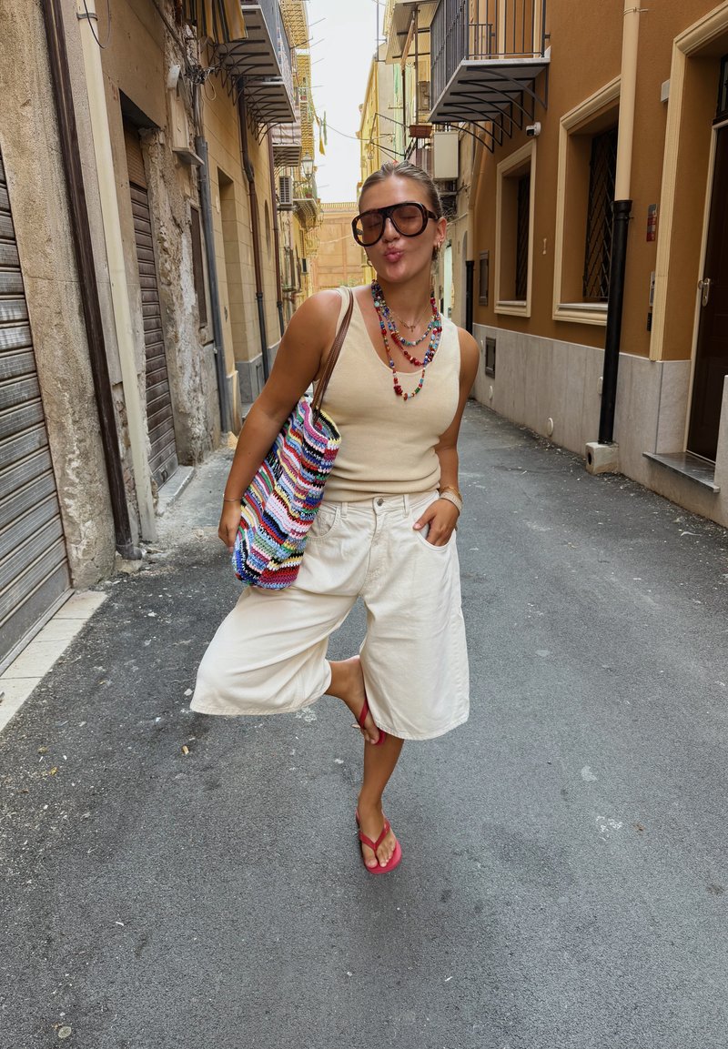Jeune femme portant des lunettes de soleil, un débardeur beige, un short blanc et des sandales rouges pose dans une rue étroite avec un sac crochet coloré et des colliers.