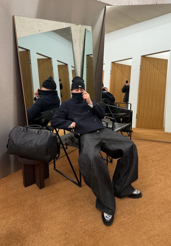 Hombre con gorro negro, chaqueta azul marino y pantalones anchos grises sentado en una silla negra junto a grandes espejos inclinados en una habitación con puertas de madera.