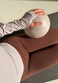 Personne en leggings marron et haut beige à manches longues tenant un ballon de gymnastique clair tout en étant assise sur un tapis.