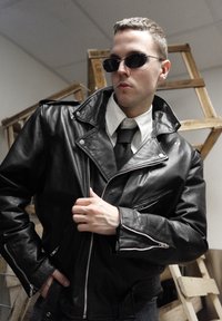 Veste de moto en cuir noir avec zips, accompagnée d'une chemise blanche et d'une cravate. Le modèle porte des lunettes de soleil noires, posant devant un fond en bois.