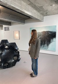 Black sculptural object with cutouts and holes, grey wall, blue abstract painting, concrete ceiling, person in a grey blazer and denim jeans.