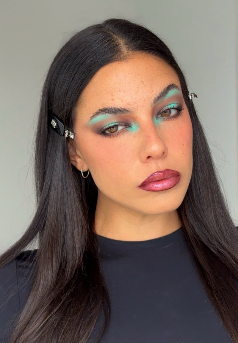 Mujer con cabello largo y oscuro, sombra de ojos turquesa, labios rosados y brillantes, pendiente de aro plateado y clips para el cabello negros, vistiendo una blusa negra sobre un fondo neutro.