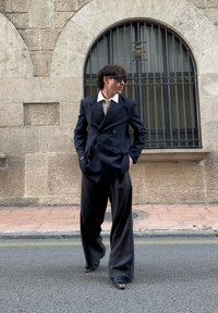 Juodas dvieilis dryžuotas dvieilis švarkas su plačiais atlapais, chaki spalvos marškiniai ir plačios juodos kelnės. Modelis stovi gatvėje šalia akmeninių sienų.