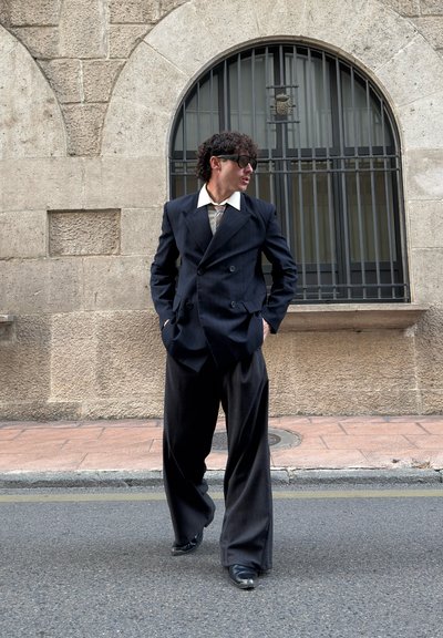 Chaqueta de traje cruzado de rayas negras con solapas grandes, camisa caqui y pantalones anchos negros. El modelo está de pie en una calle con muros de piedra.