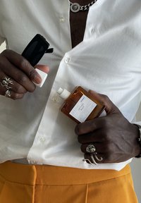 Person in white shirt and orange pants holding a bottle of Vetiver Root cologne and black sunglasses, wearing silver rings and chain necklace.