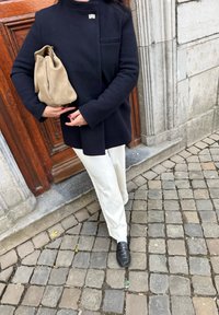 Czarny wełniany płaszcz z małym metalowym akcentem, w połączeniu z kremowymi spodniami i torebką z brązowego zamszu, na tle brukowanej ulicy.