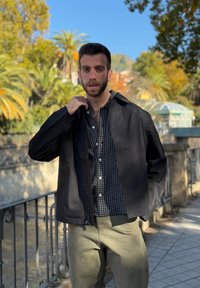 Άνδρας με κοντά σκούρα μαλλιά και γενειάδα, ντυμένος με μαύρο μπουφάν και καρό πουκάμισο, στέκεται σε εξωτερικό χώρο δίπλα σε κιγκλίδωμα με δέντρα και γαλανό ουρανό στο βάθος.