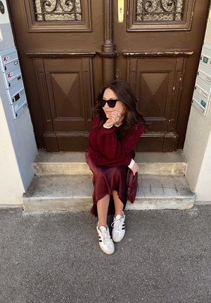 Weinroter Pullover über einem weißen Hemd, weinroter Satinrock und weiße Turnschuhe mit kastanienbraunen Streifen. Sitzend auf Steinstufen neben einer braunen Tür.