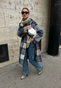 Dugi karirani kaput u plavoj, smeđoj i bijeloj vuni, bež rebrasti rolak, plave traperice s prednjim prorezima i bež cipele s oštrim vrhom. Bijela torba.