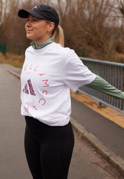 Camiseta deportiva blanca con logotipo circular rojo y detalles negros, combinada con leggings negros. El fondo muestra un camino y vegetación.