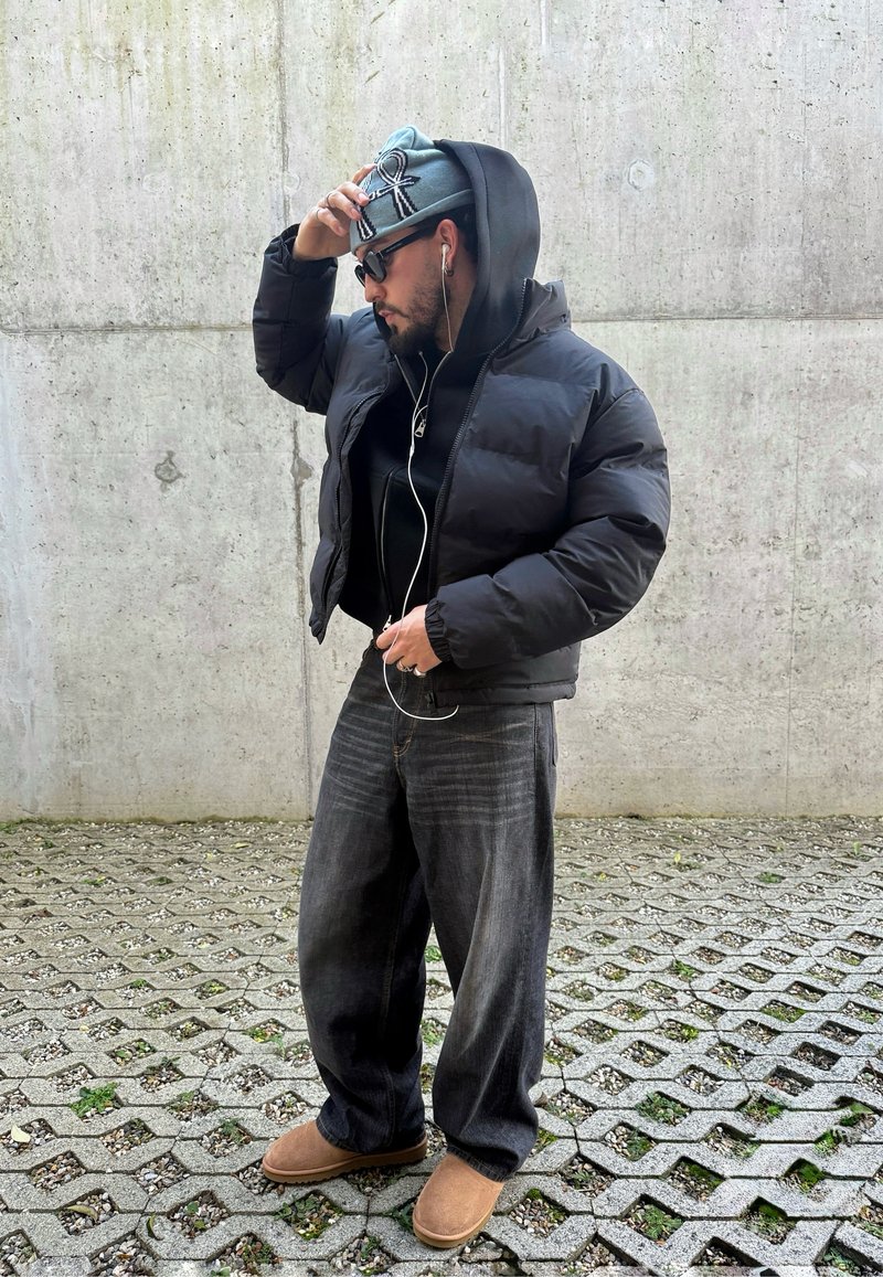 Veste matelassée noire, sweat à capuche noir en dessous, bonnet bleu avec motif, jean en denim foncé, chaussures à enfiler couleur fauve, casque audio et lunettes de soleil.