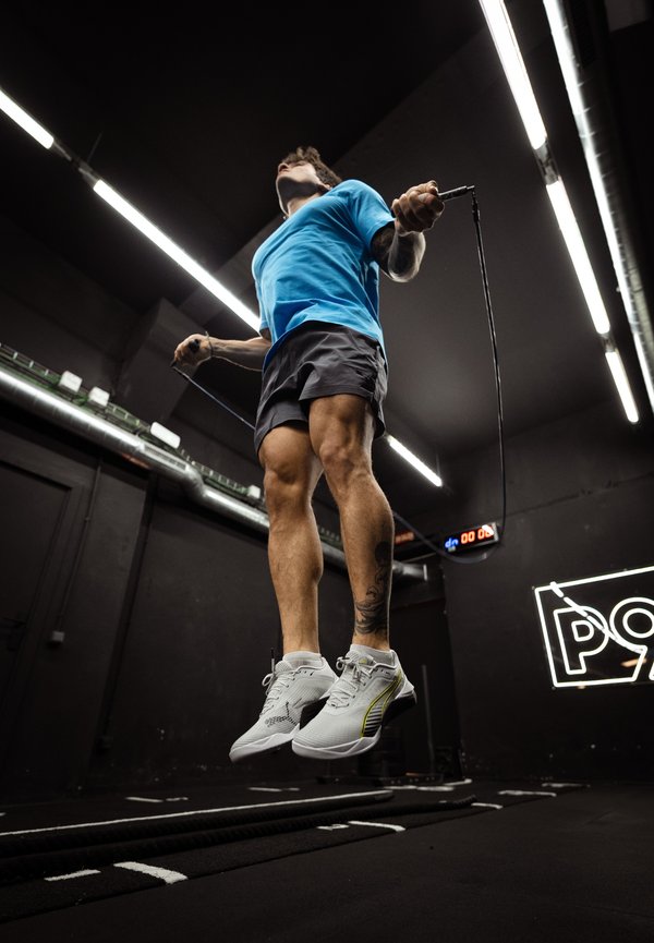 Mann springt Seil im Fitnessraum, trägt blaues Shirt, schwarze Shorts, weisse Sneakers, mit sichtbarem Bein-Tattoo und dunklen Deckenleuchten darüber.