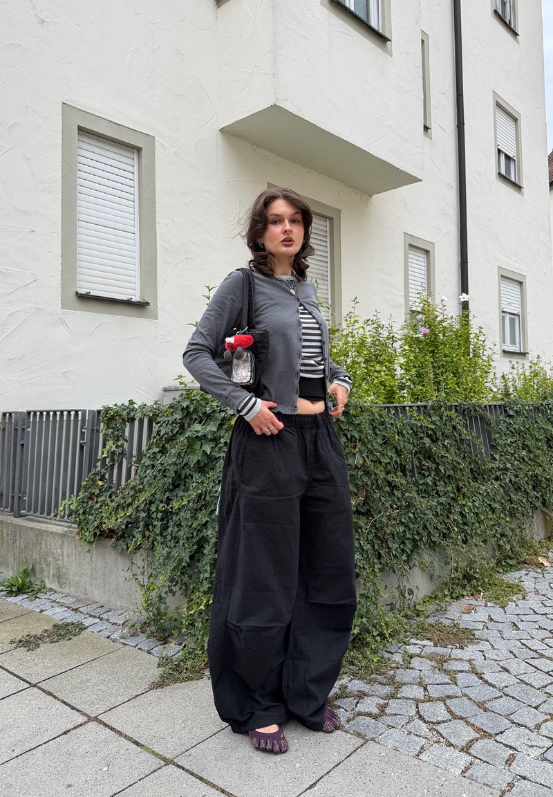 Joven mujer de pie en la acera frente a un edificio blanco con ventanas cerradas, vistiendo pantalones negros holgados y una chaqueta gris corta.