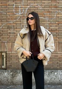 Jachetă bej din lână cu accente maro, pulover gros tricotat maro închis, pantaloni negri croiți și o geantă mică neagră. Ochelari de soare simpli.