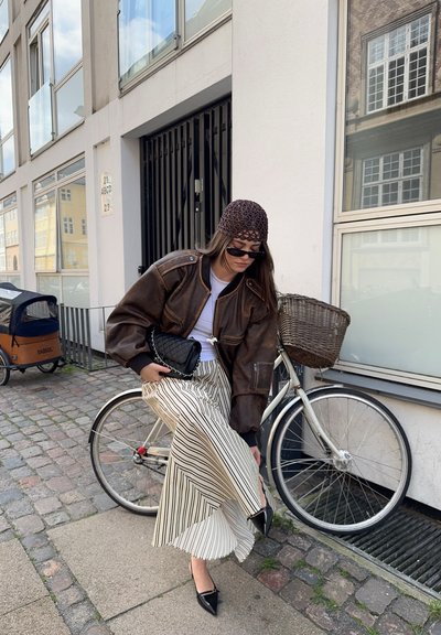Chaqueta bomber de cuero marrón, camiseta blanca, falda rayada y zapatos planos negros con puntera. Los accesorios incluyen un bolso negro y gafas de sol.