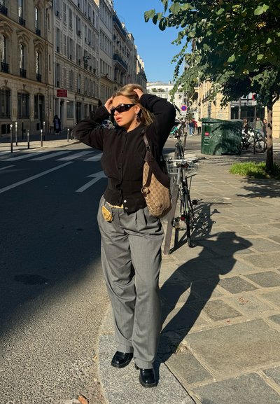 Camisa negra de botones, pantalones gris de tiro alto y zapatos negros. Lleva una bolsa de hombro marrón con estampado. Día soleado y brillante, entorno urbano.