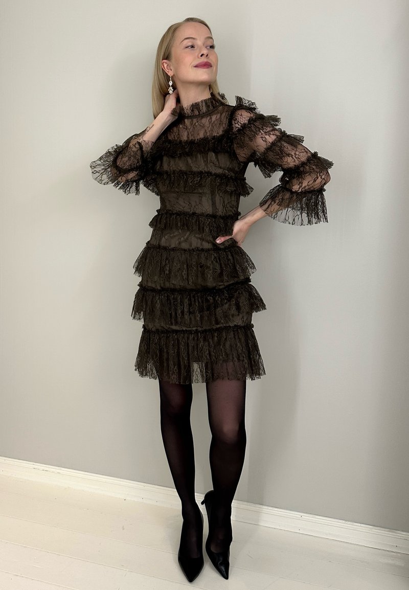Lace dress in dark brown with layered frills, sheer long sleeves, fitted waist, and a flared skirt. Black high heels complete the look.