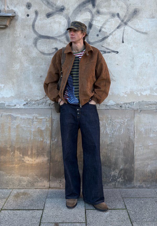 Brown suede jacket, green and blue striped shirt, wide-leg dark denim jeans, beige boots, and camouflage cap. Graffiti-covered wall in the background.