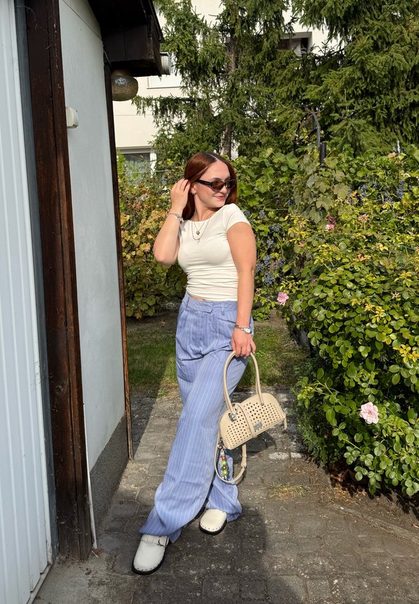 Mujer con gafas de sol, con top blanco y pantalones azules a rayas, sosteniendo un bolso beige de pie en un camino del jardín cerca de la vegetación.