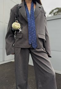 Personne portant un costume gris surdimensionné avec une cravate bleue à motif, portant un sac noir orné d'un porte-clés peluche rond en plein air.