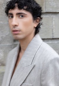 Joven con cabello rizado oscuro que lleva un blazer gris claro, de pie frente a una pared de ladrillos de piedra, mirando ligeramente hacia un lado.