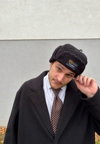 Svart, texturerad fleecehatt med vikta öronlappar och en logotypärm. Bärs tillsammans med en svart kappa och en mönstrad slips, mot en grå vägg.