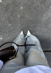 Hvide spidse hæle, lyseblå posede stribede jeans, sort lædertaske og en hvid skjorte mod en grå belægning.
